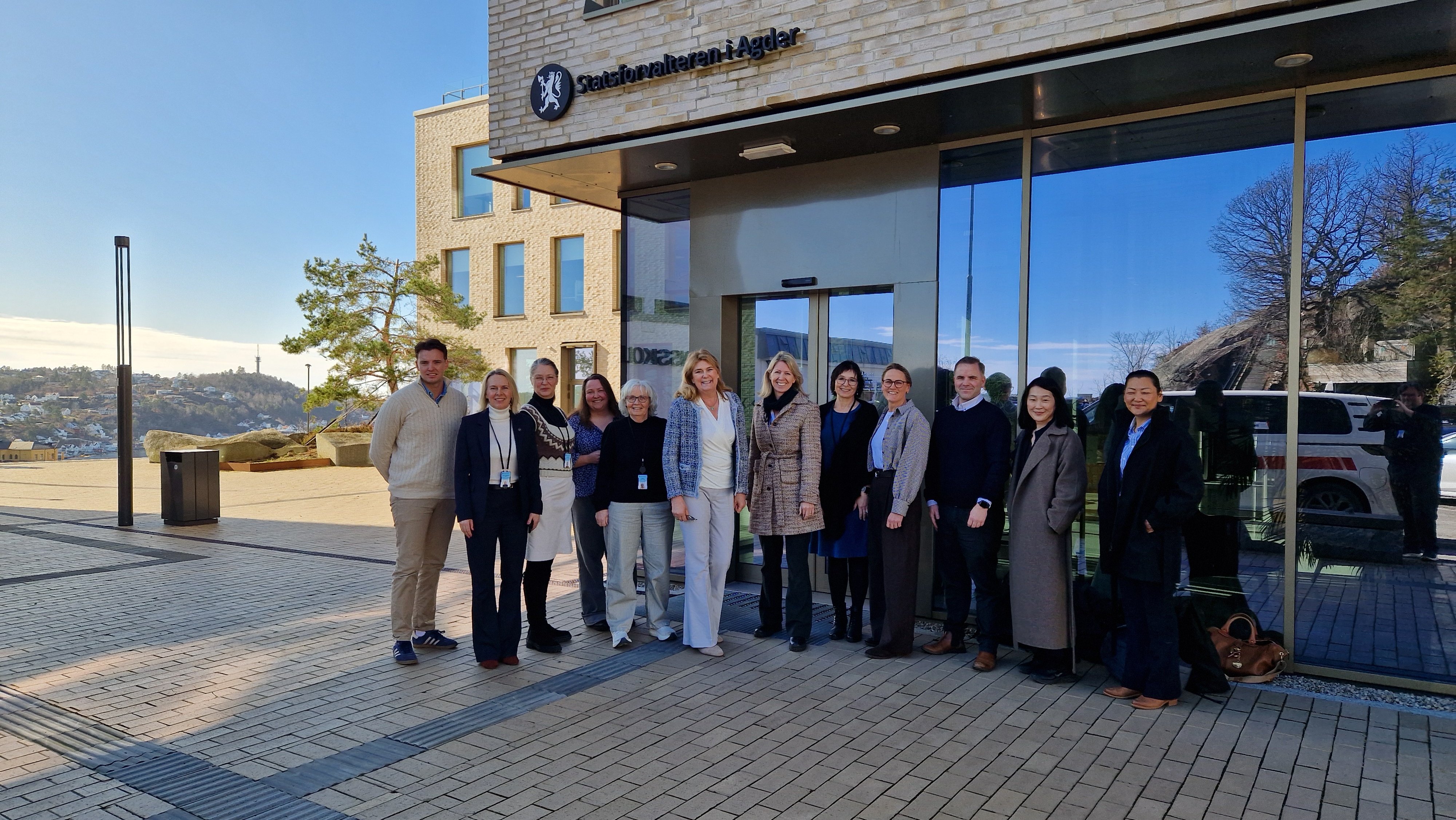 En gruppe smilende mennesker står på rekke fremfor bygg med logoen "Statsforvalteren i Agder"