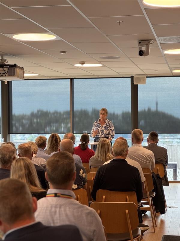Ein person held innlegg frå talarstolen i eit møterom. Publikum sit på stolrekkjer og lyttar, med store vindauge og utsyn i bakgrunnen.