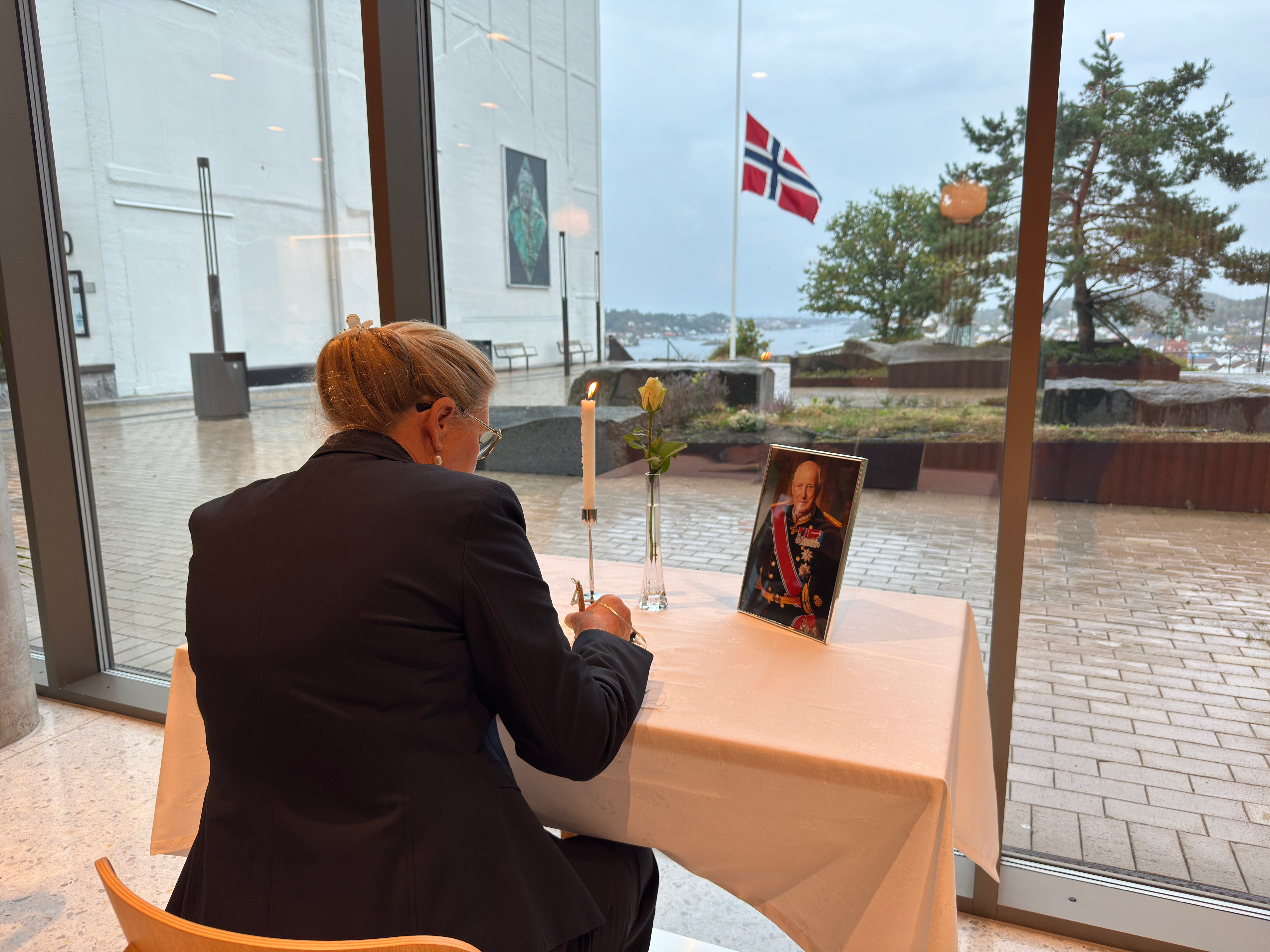 En person skriver i kondolanseprotokollen som er lagt frem ved Statsforvalterens kontor på Fløyheia i Arendal. På bordet står et tent lys, en hvit rose og et innrammet portrett av Kong Harald V, mens det norske flagget er heist på halv stang utenfor.