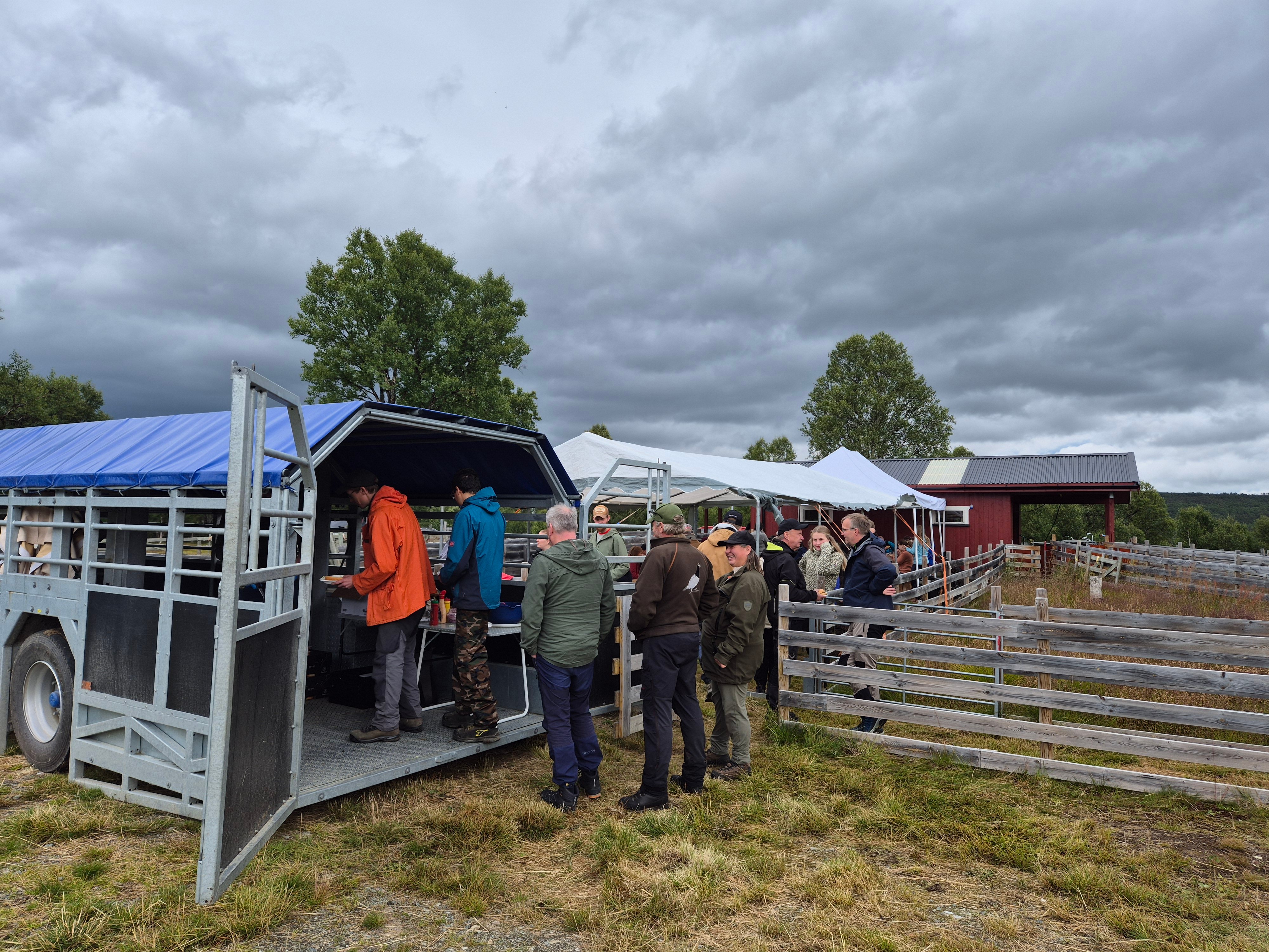 En rekke personer som står i kø inn i en stor, åpen tilhenger med matservering, ved siden av en innhegning av plankegjerde og et rødt bygg i bakgrunnen.