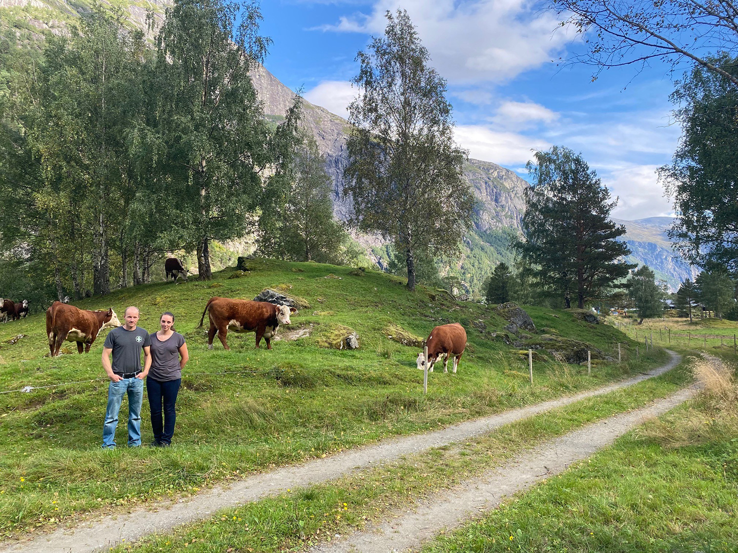 Marius Remmem og Linn Kristin Hestad står ved ein traktorveg. Bak dei går fleire kyr og beiter på grønt gras. Tre og fjell i bakgrunnen.