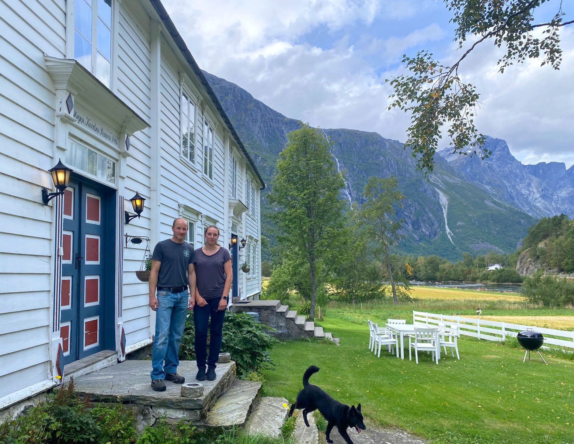 Marius Remmem og Linn Kristin Hestad står på trappa framfor eit kvitt våningshus. En svart hund går ned trappa. Fjell i bakgrunnen og grønt gras ved huset.