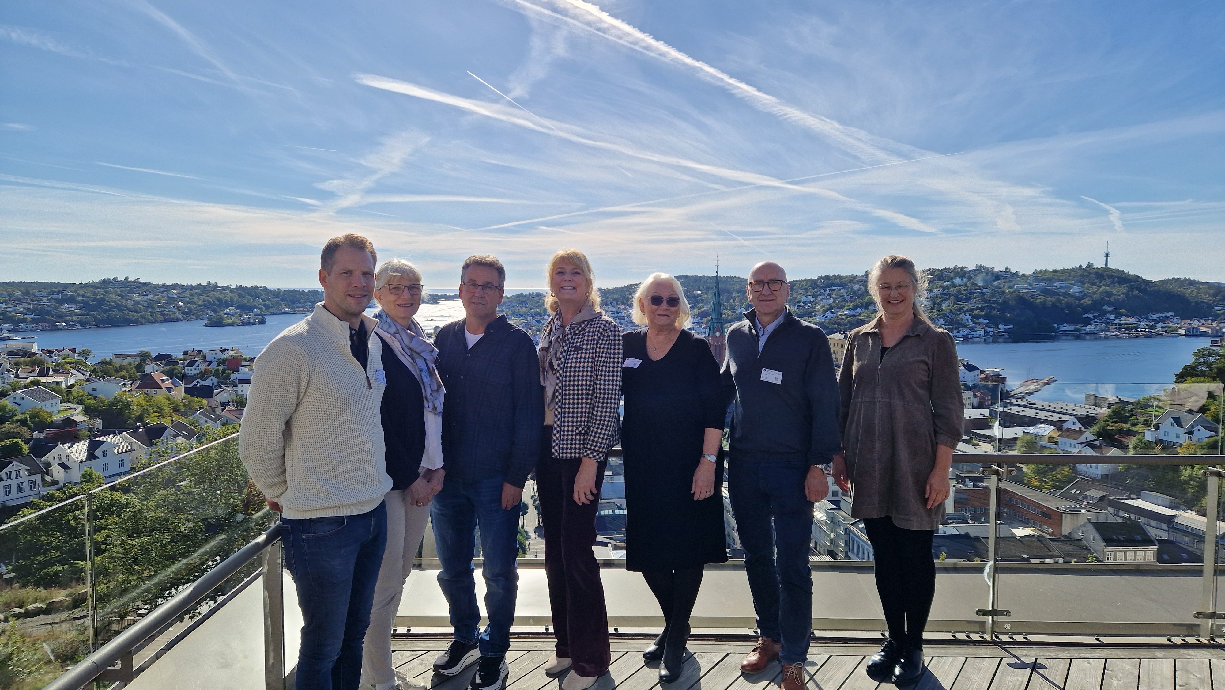 Syv mennesker står på linje på en terrasse med Arendal by i bakgrunnen