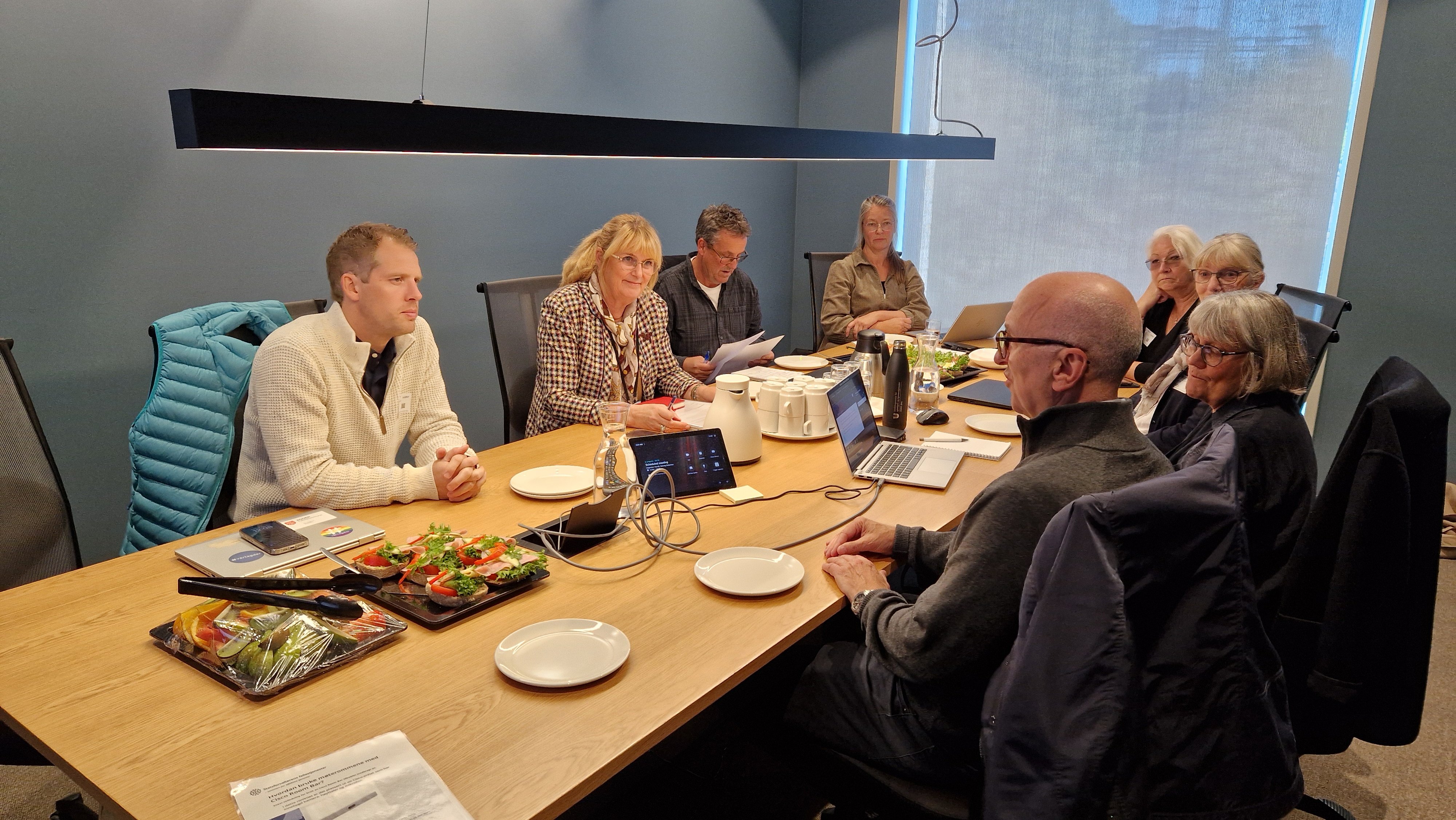 Gruppe mennesker rundt møtebord, med PC-er, kaffekanne og papirer