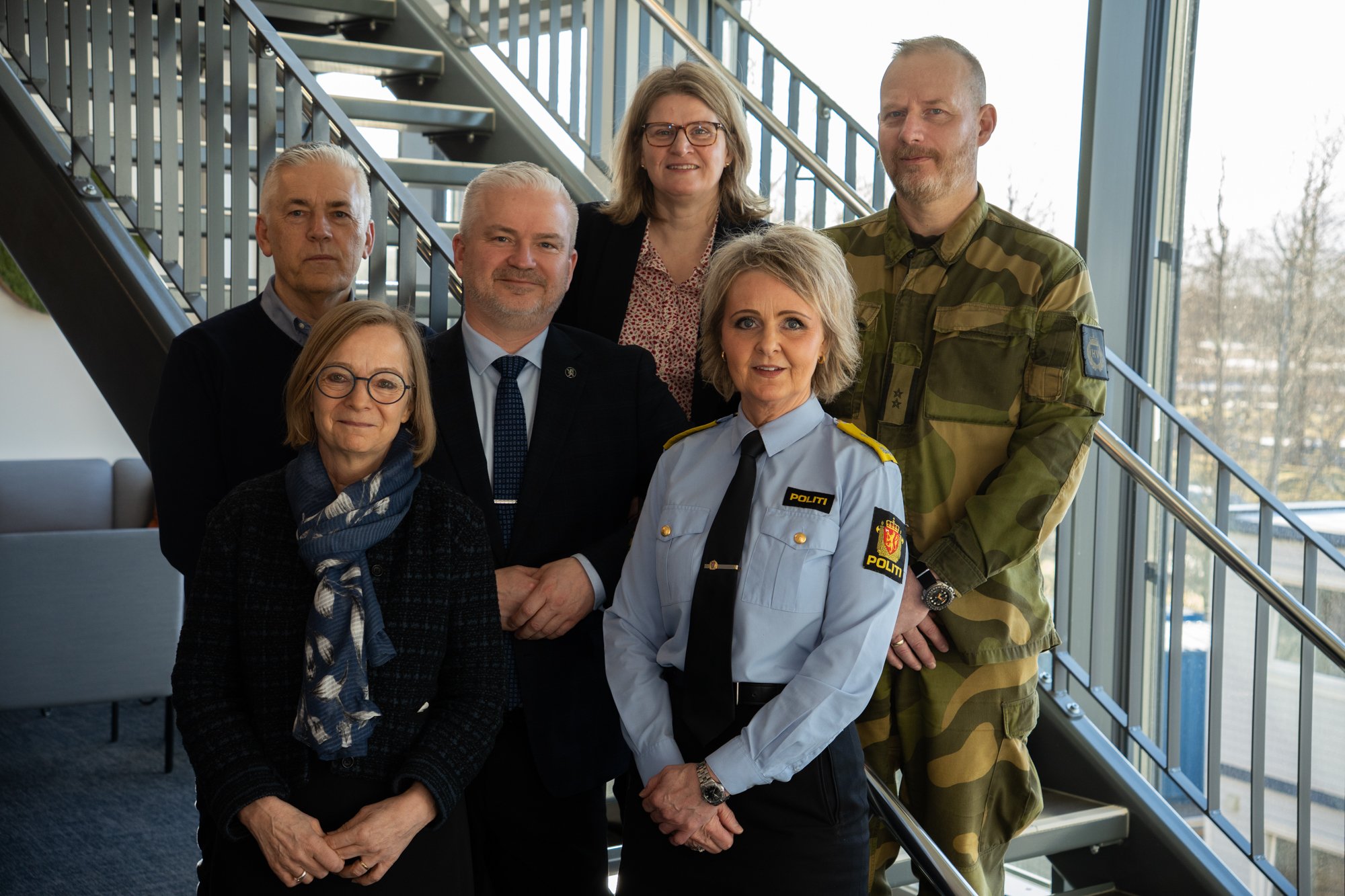 Seks personer står oppstilt ved en trapp med vinduer i bakgrunnen.