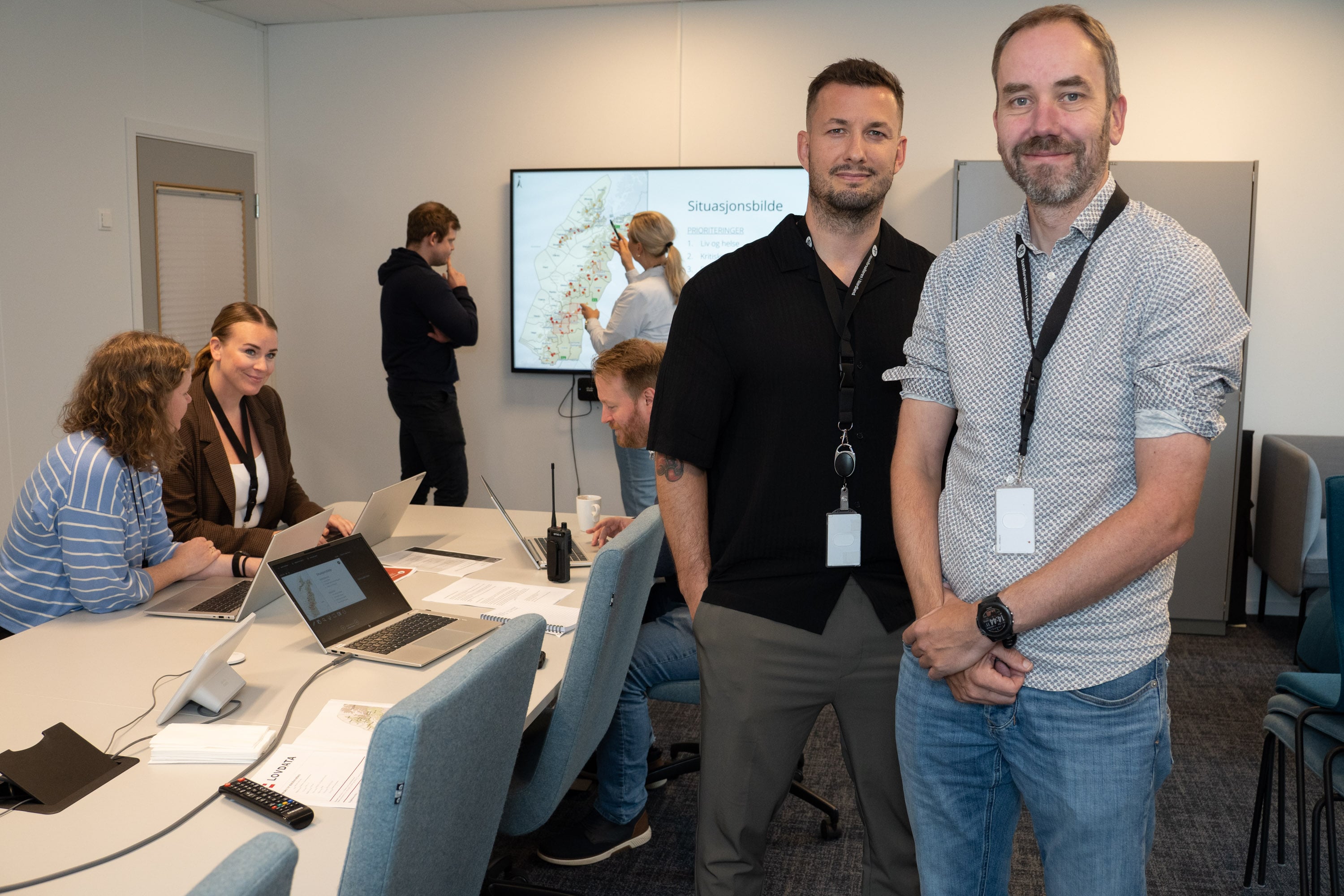 To personer står ved et møtebord. I bakgrunnen ser man personer som sitter foran PC-er ved bordet og to personer som står ved en TV-skjerm på veggen.