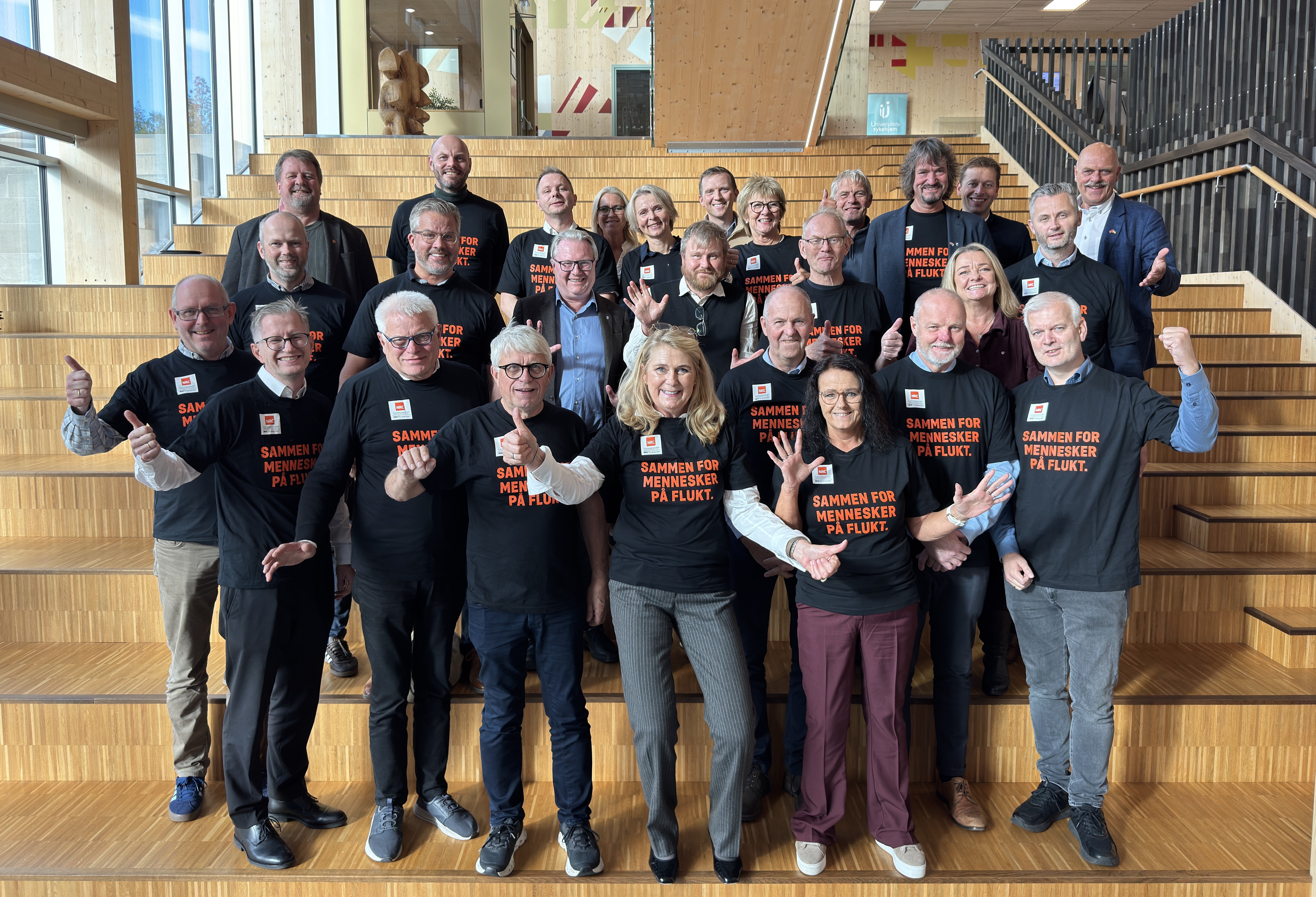 Gruppebilde fra Ordfører- og kommunedirektørkonferansen 2026. Om lag 25 deltakere står samlet i en tretrapp inne i et moderne konferansebygg med store vinduer og treinteriør. Statsforvalteren i Agder, fylkesordføreren og ordførerne i Agders kommuner er avbildet sammen. Flere har på seg svarte T-skjorter med teksten «Sammen for mennesker på flukt».