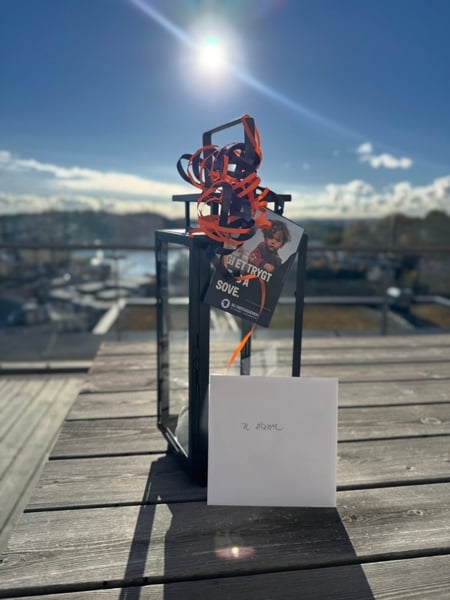 Ei svart lykt pynta med oransje band står på eit trebord utandørs. Framfor lykta står eit kort merka «Til Åseral». Ei brosjyre frå TV-aksjonen er festa til lykta, og i bakgrunnen er det utsikt mot sjø og busetnad i sollys.