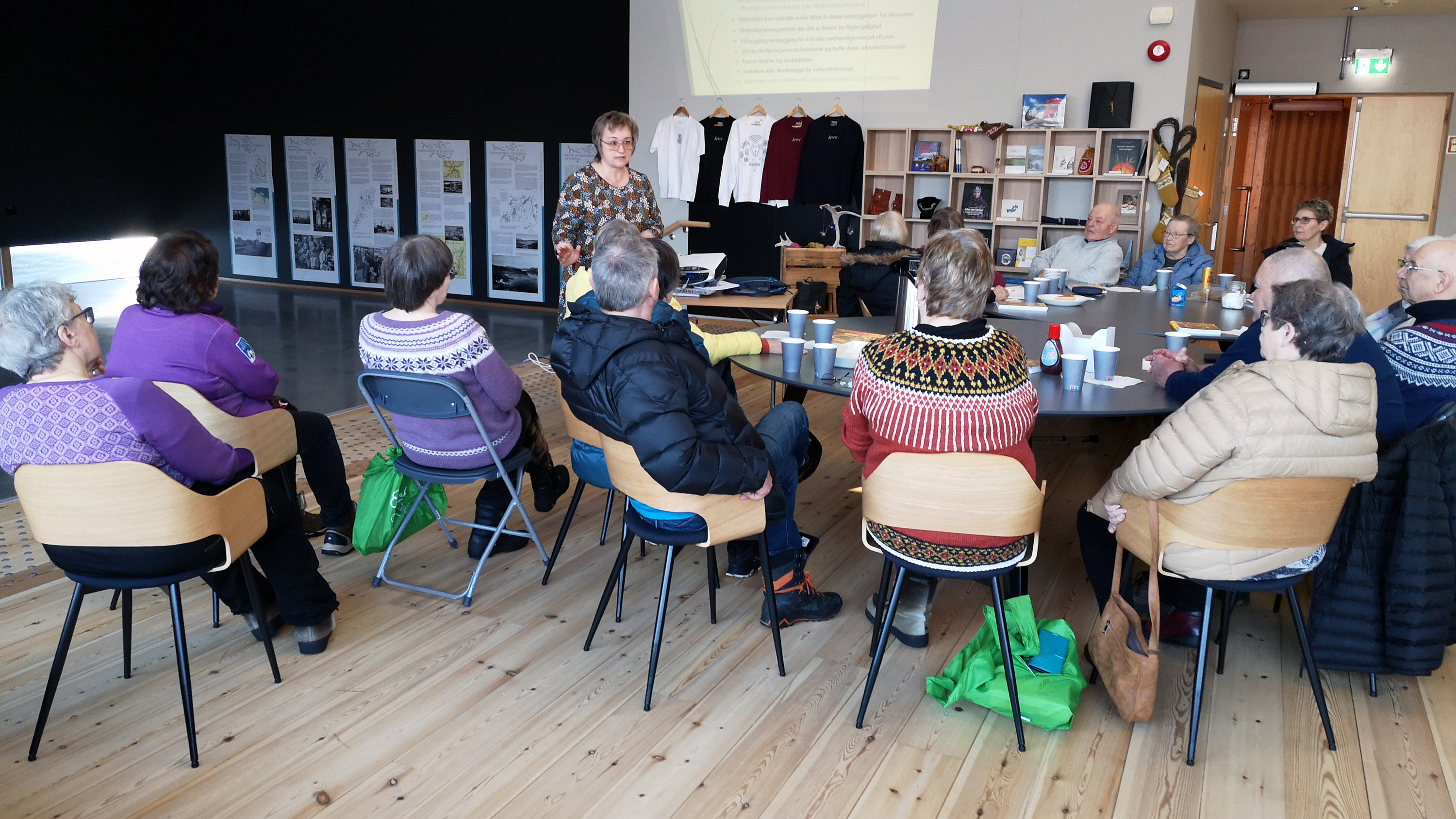 Undersøkelsen og tilskuddsordninger presenteres for pensjonistforeningen i Neiden i Sør-Varanger. Foto: Åsmund Røst. Mennesker sitter på stoler rundt bord og lytter til kvinne som står og holder en presentasjon.