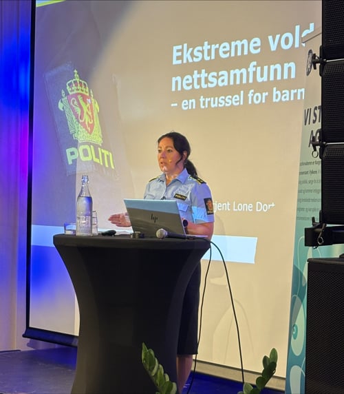 Politioverbetjent Lone Austad fra Kripos står på scenen under et seminar og holder en presentasjon. På storskjermen bak vises lysbildet «Ekstreme voldelige nettsamfunn – en trussel for barn og unge» med logoen til Politiet/Kripos. Innlegget handler om risikoen ekstreme nettfellesskap utgjør for barn og unge og behovet for forebygging og samarbeid på tvers av tjenester.
