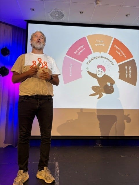 Ole Morten Mouridsen fra Stine Sofies Stiftelse holder innlegg på en scene under et seminar. På storskjermen bak vises en illustrasjon om handlingskompetanse og faktorer som kunnskap, personlige faktorer, tro på systemene, organisatoriske faktorer og verdier/barnesyn. Innlegget handler om hvordan voksne kan styrkes til å forebygge, avdekke og handle ved bekymring for vold og overgrep mot barn.