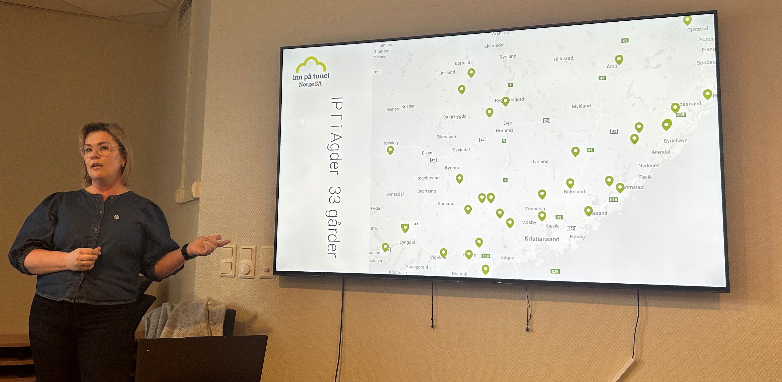 Kvinne står ved siden av skjerm med punkter på kart og teksten IPT i Agder 33 gårder. 