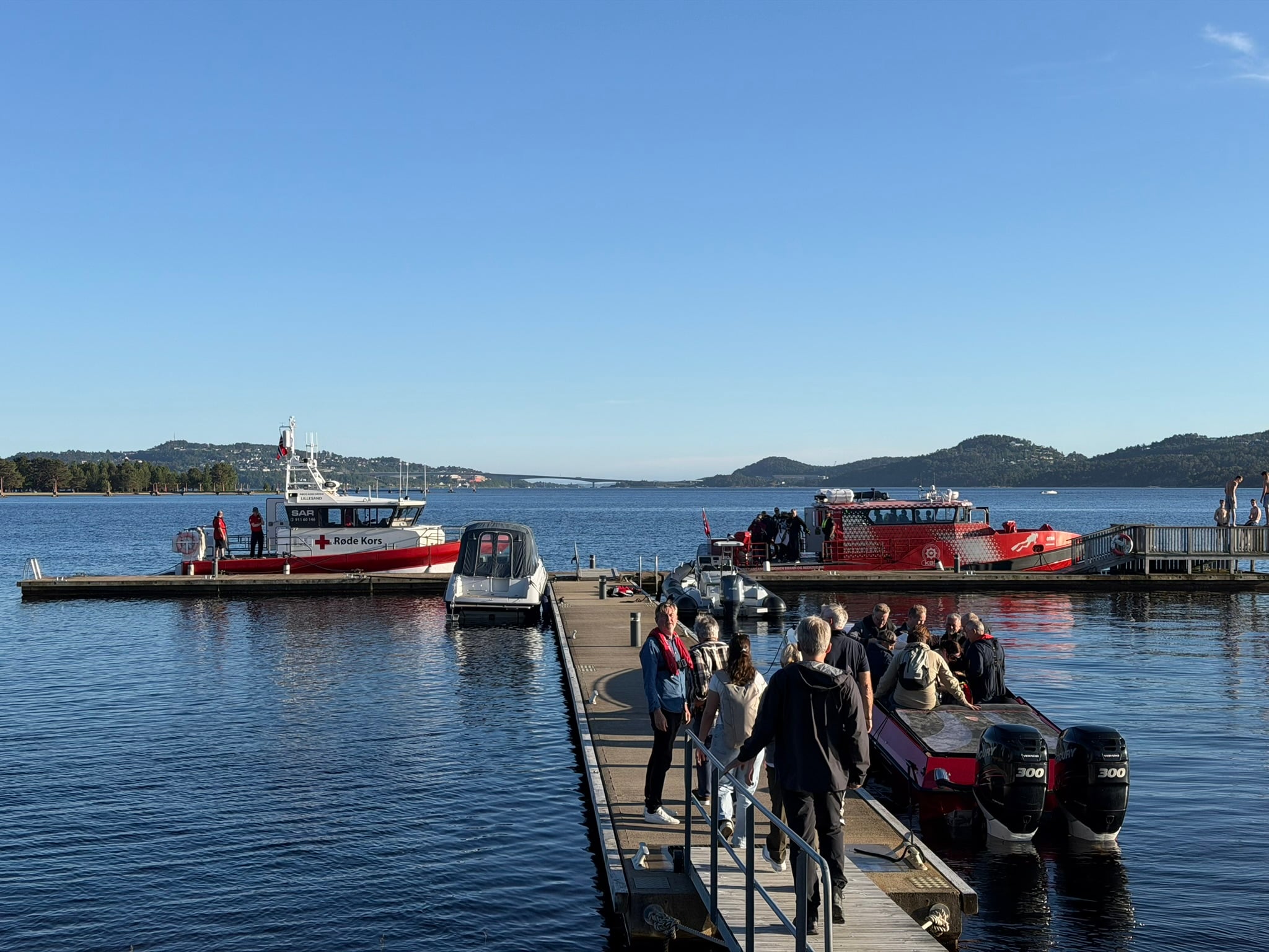 Brygge ved sjøen med flere personer som går om bord i småbåter, mens redningsbåt fra Røde Kors og en annen beredskapsbåt ligger fortøyd i rolig vann, med skjærgård og bro i bakgrunnen under klar blå himmel.