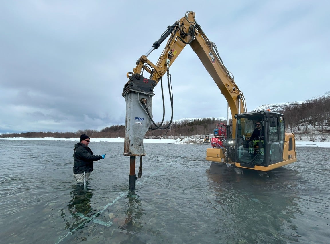 Ved hjelp av gravemaskin monteres fundamentet til flyteristfellen i Reisaelva. Selve flyteristen vil bli montert i juni. Foto: Hermann O. Hermansen, Arktisk Geotek AS. Gravemaskin og personell arbeider ute i elv.