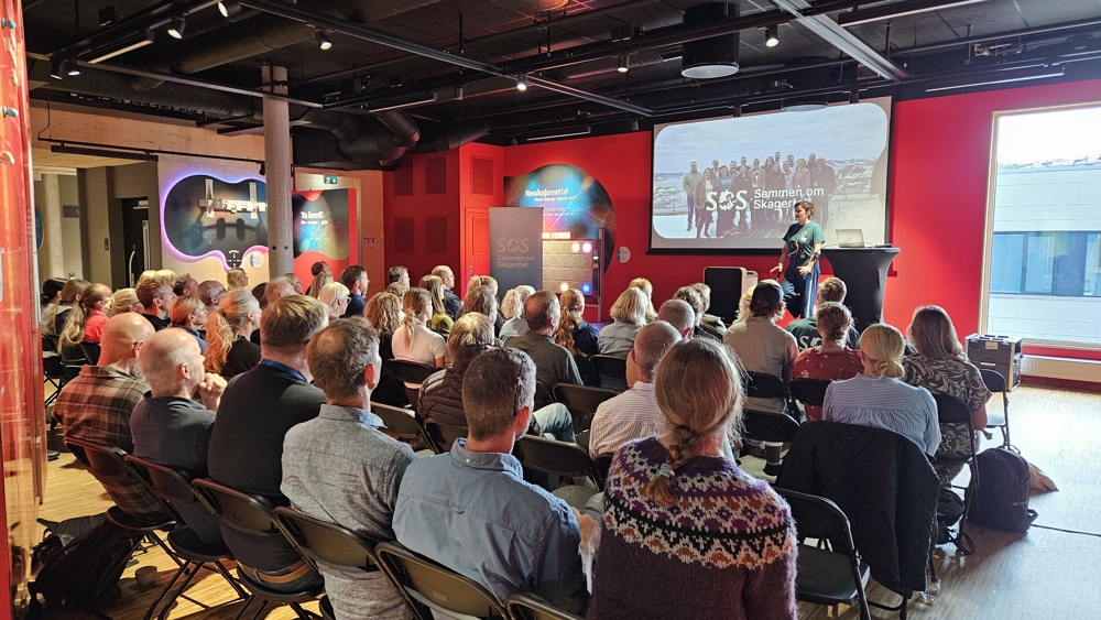 Deltakere sitter i et konferanselokale og følger åpningen av workshop for SOS-Saman om Skagerrak. En person holder innlegg fra scenen foran en storskjerm som viser programlogoen og et bilde fra tidligere aktiviteter i programmet. Lokalet er fylt med deltakere vendt mot scenen.