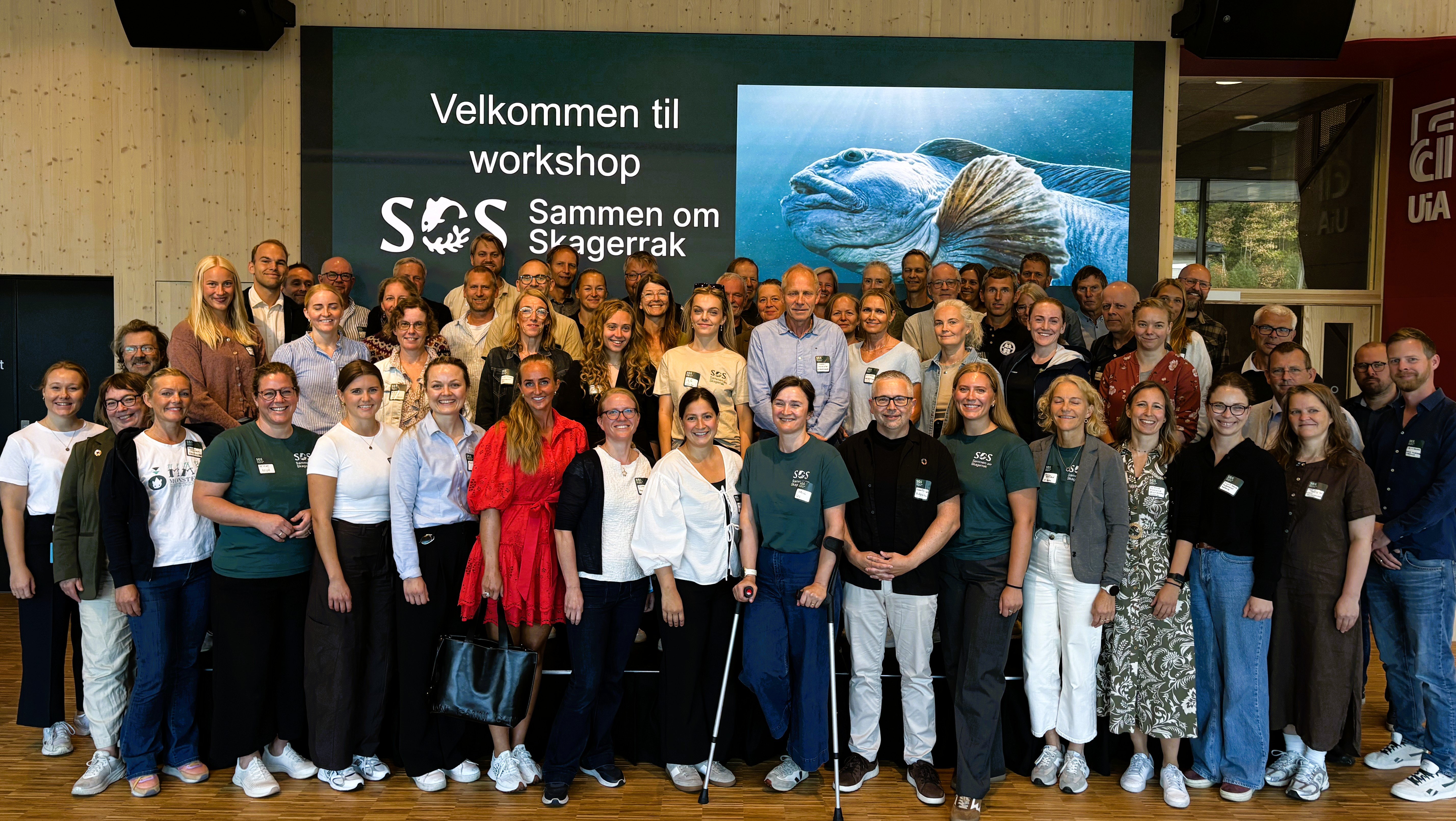 Gruppebilete av om lag 60 deltakarar på workshop for SOS-Saman om Skagerrak. Deltakarane står samla framfor ein stor skjerm med programlogo og teksten «Velkommen til workshop SOS Saman om Skagerrak». Biletet er teke innandørs i eit konferanselokale ved Universitetet i Agder.
