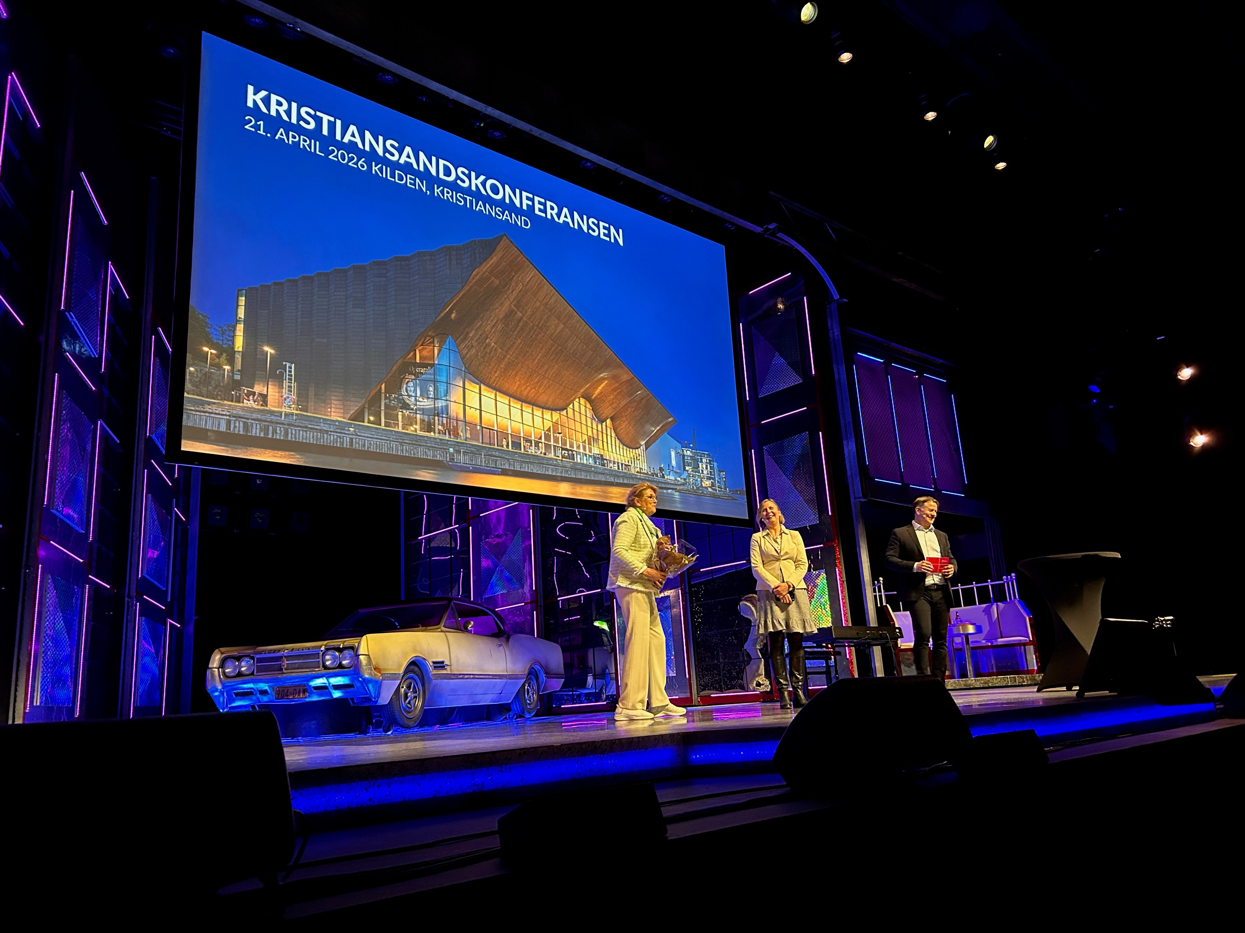 Bilde av smilende mennesker på scene, en av dem med blomster i hendene.