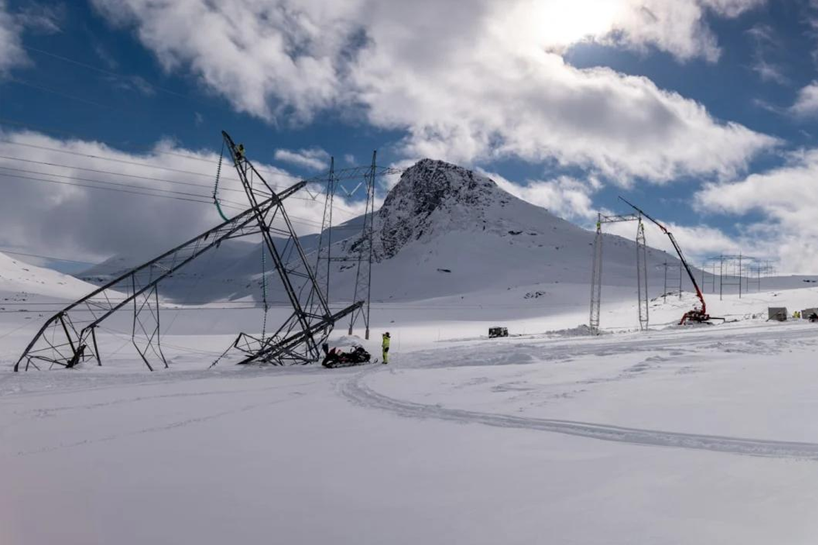 Viser en knekt mast på et fjell som er nedsnødd. En person står i nærheten av masten og arbeider. 