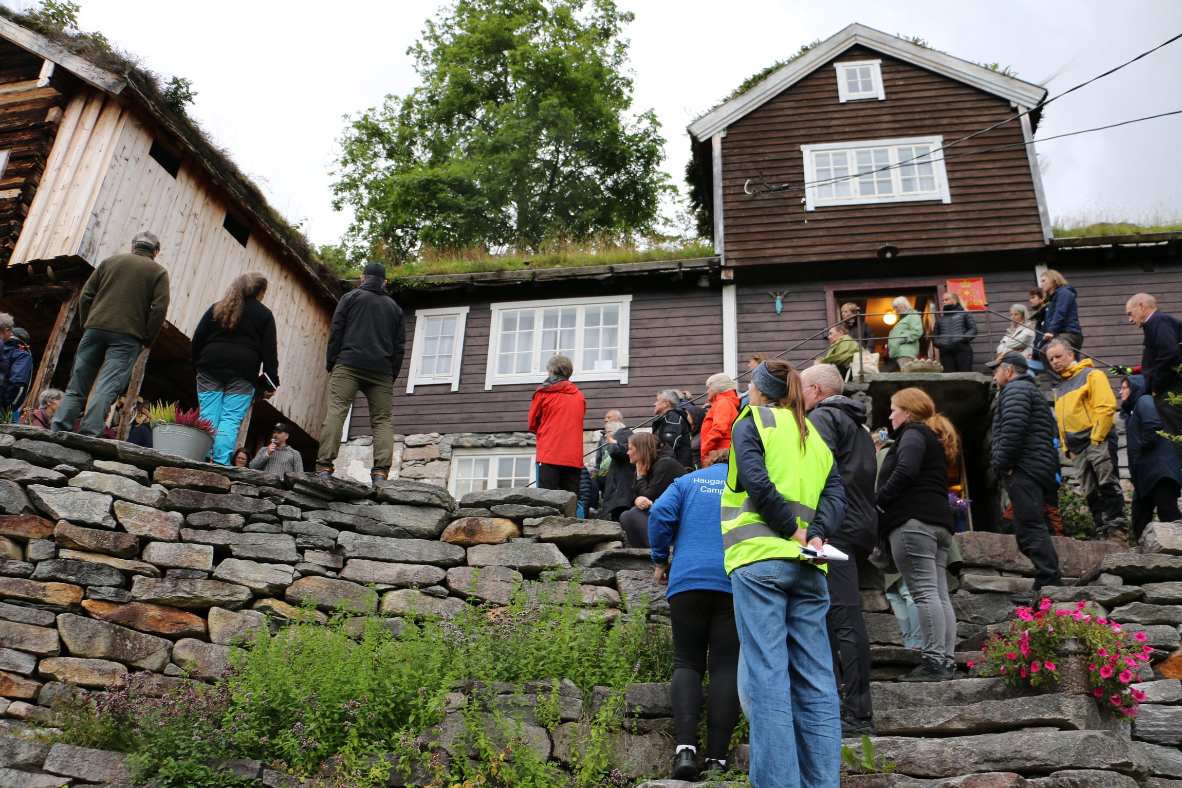 Ei gruppe personar står framfor eit gardshus. En mann med mikrofon snakkar til gruppa.