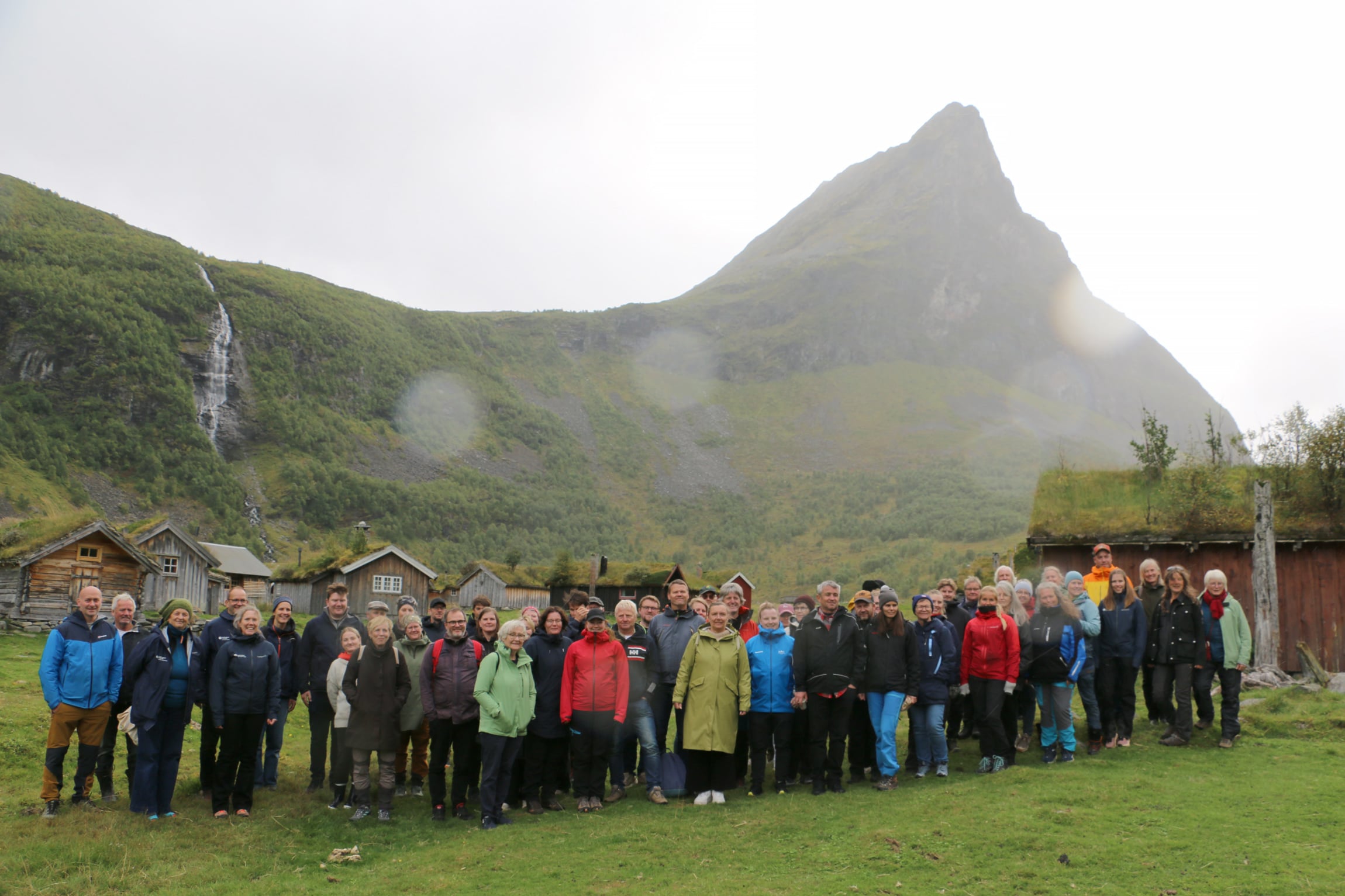 Ei stor gruppe menneske ser mot kamera. Dei står på ei seter i med fjell i bakgrunnen.