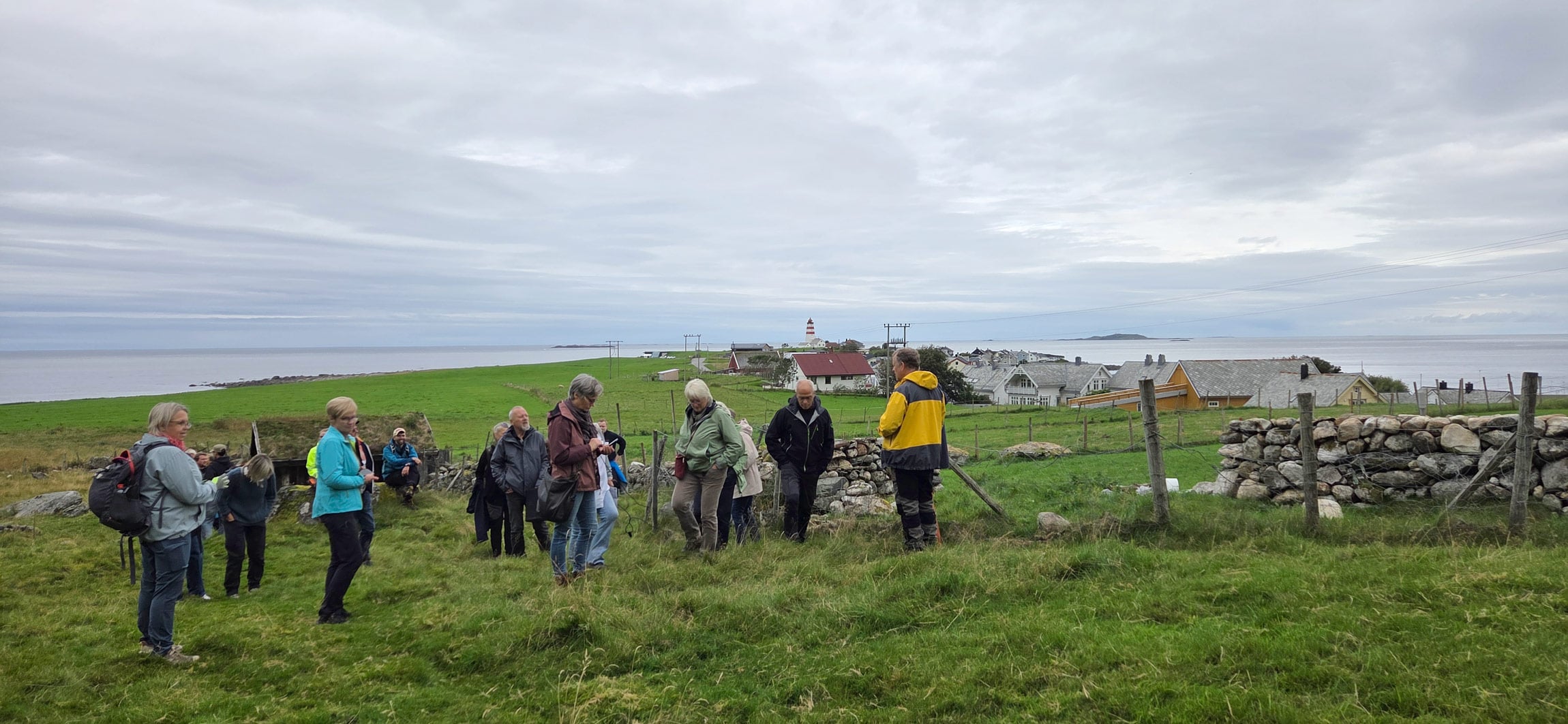 Ei gruppe personar står på ei mark i eit kystlandskap. I bakgrunnen er det fleire hus, eit fyrtårn og havet.