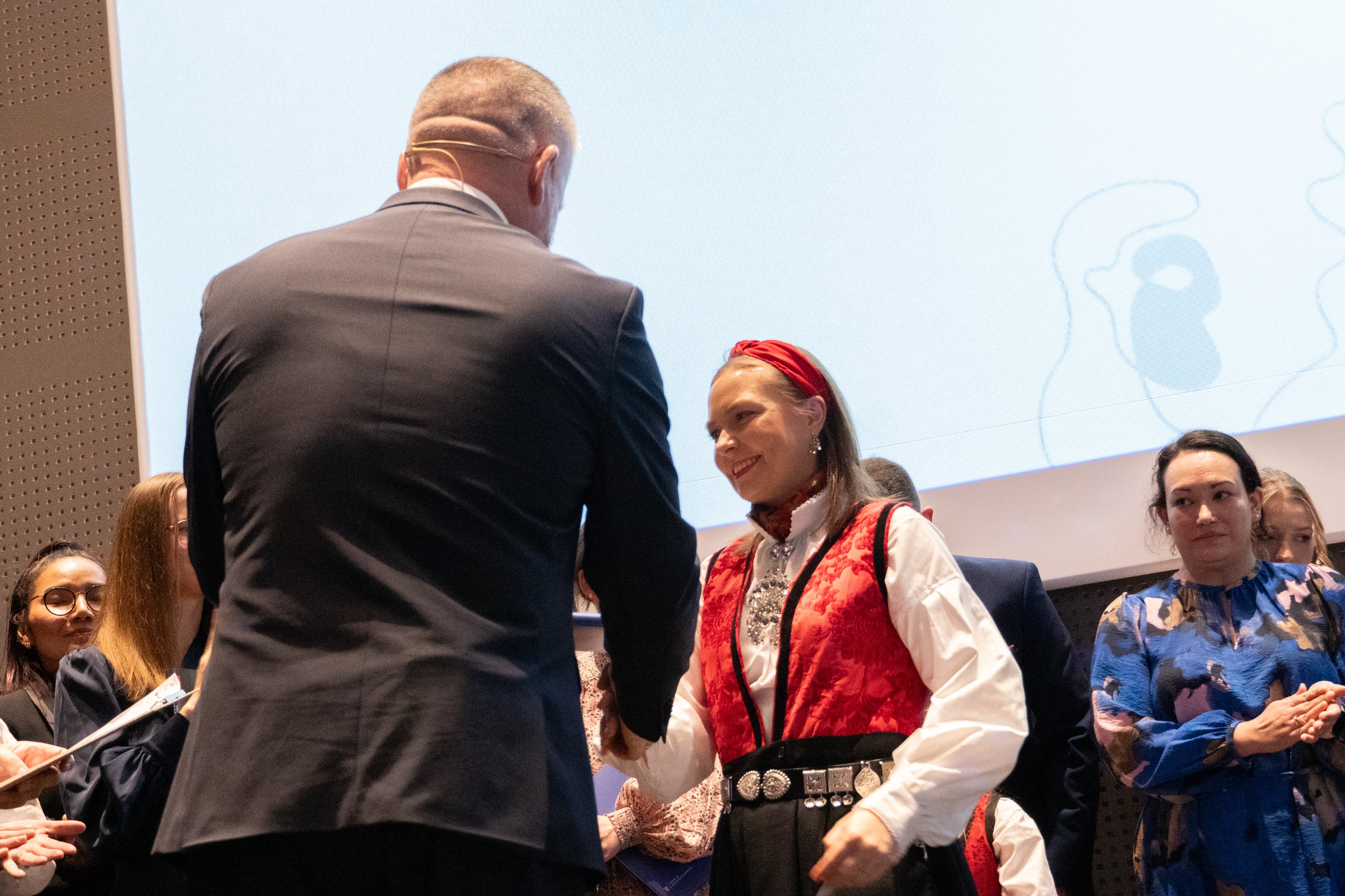 Kvinne står på en scene og mottar en bok fra en mann med pyntede mennesker i bakgrunnen.