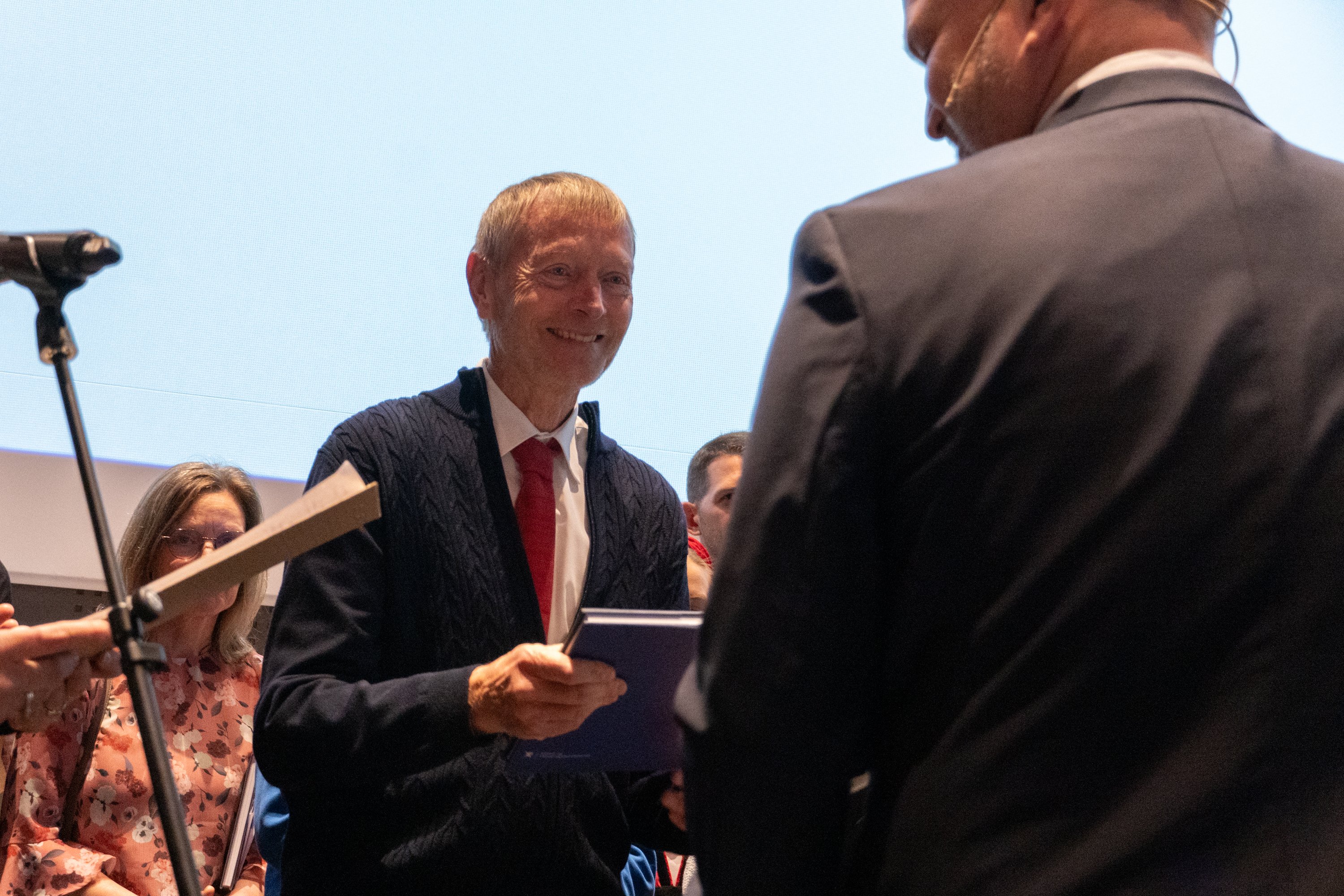 Mann står på en scene og mottar en bok fra en mann med pyntede mennesker i bakgrunnen.
