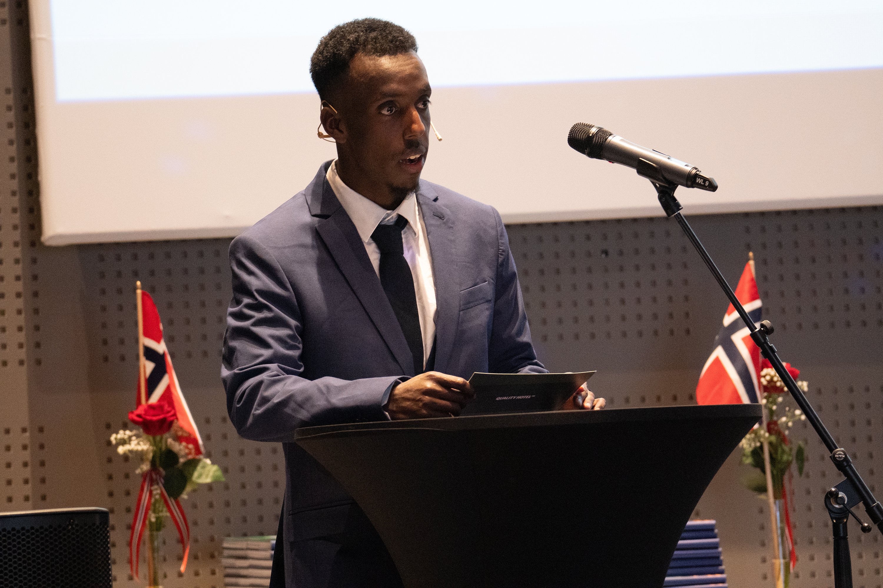 Mohamed Jirde Ali var gjestetaler under årets seremoni. Foto: Emil Harjo Indsetviken / Statsforvalteren i Nordland. En mann i dress står bak en talestol med norske flagg i bakgrunnen.