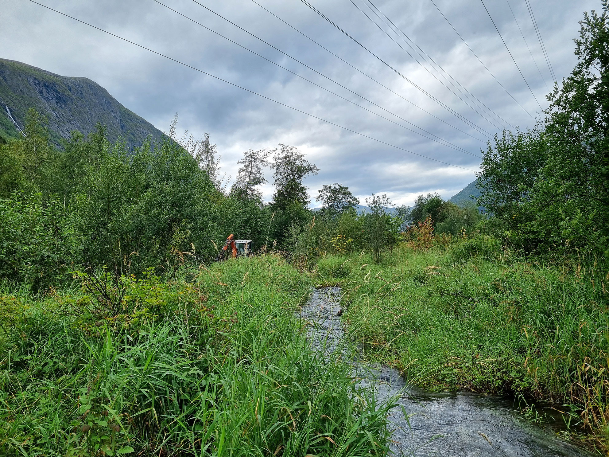 Ei smal elv med mykje gras langs elvekantane. Ei gravemaskin i bakgrunnen.