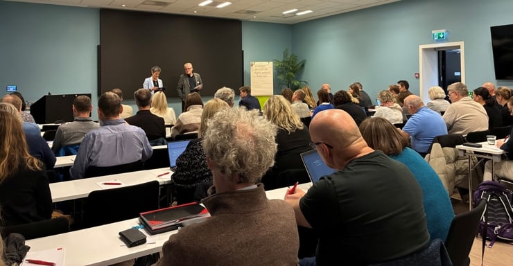 Ei større forsamling av personar sett bakifrå på eit konferanserom, med to personar som står fremst i rommet og presenterer.