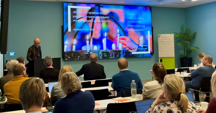 Ei gruppe personar sett bakifrå i eit konferanserom, men ein person som står fremst i rommet og presenterer med ein stor skjerm på veggen.
