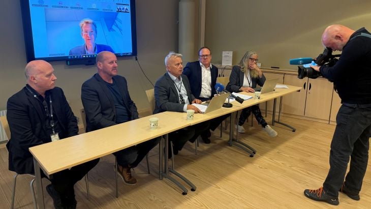 Seks mennesker som sitter ved et bord på en pressekonferanse. En mann med kamera står forran. På veggen bak ser vi møtedeltagere som er med digitalt. 