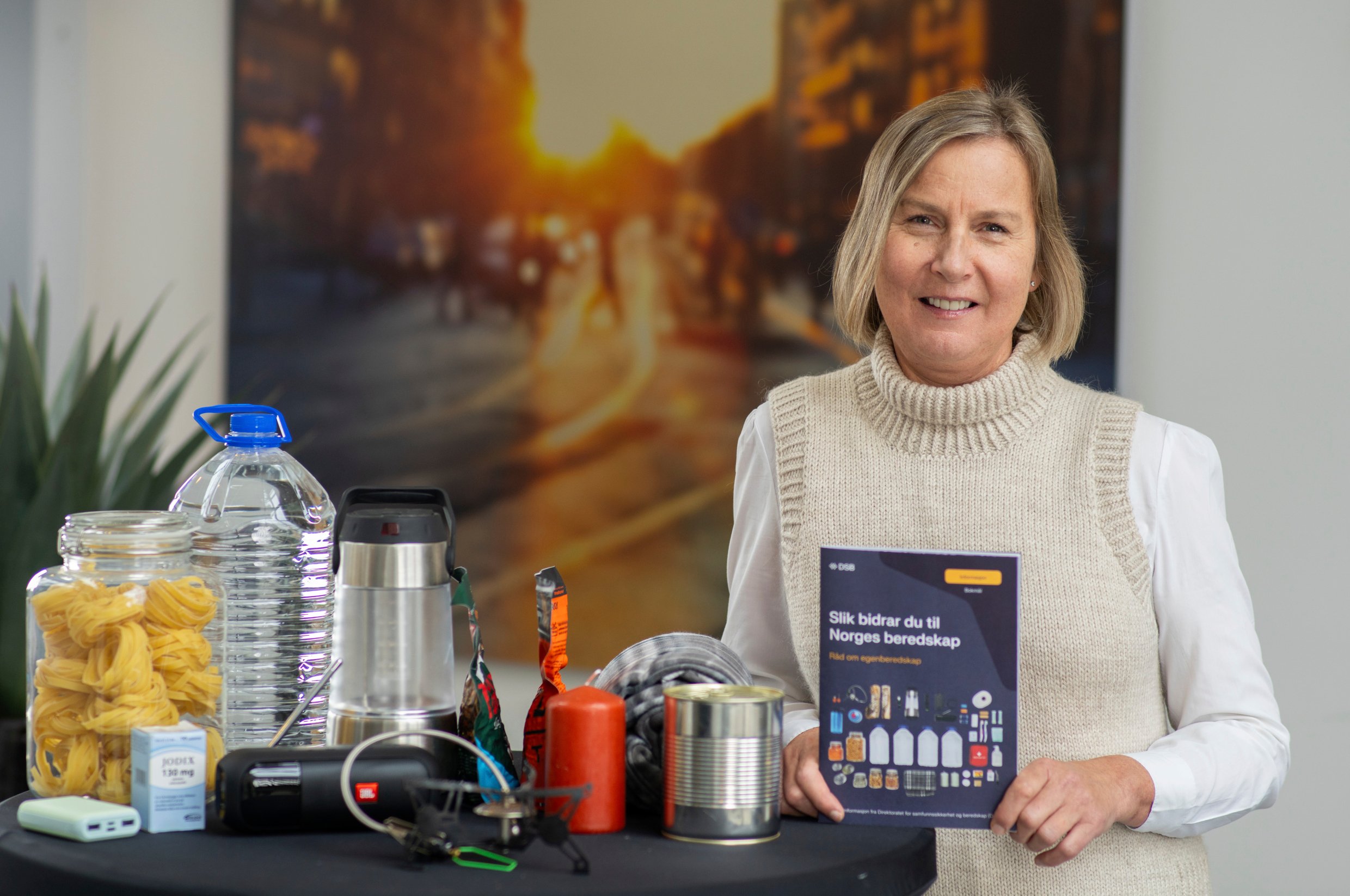 Bilde av Elisabeth Aarsæther som holder brosjyre med tittelen "Slik bidrar du til Norges beredskap". Til venstre et bord med beredskapsutstyr som vann, powerbank mv.