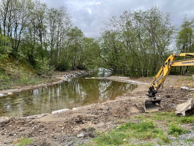 Gul gravemaskin til høyre i bildet som graver i et vassdrag. 