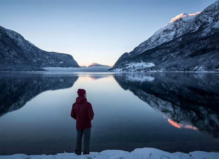 Ein person med raud lue står med ryggen til fotografen og ser ut over eit fjordlandskap med fjell på begge sider av fjorden