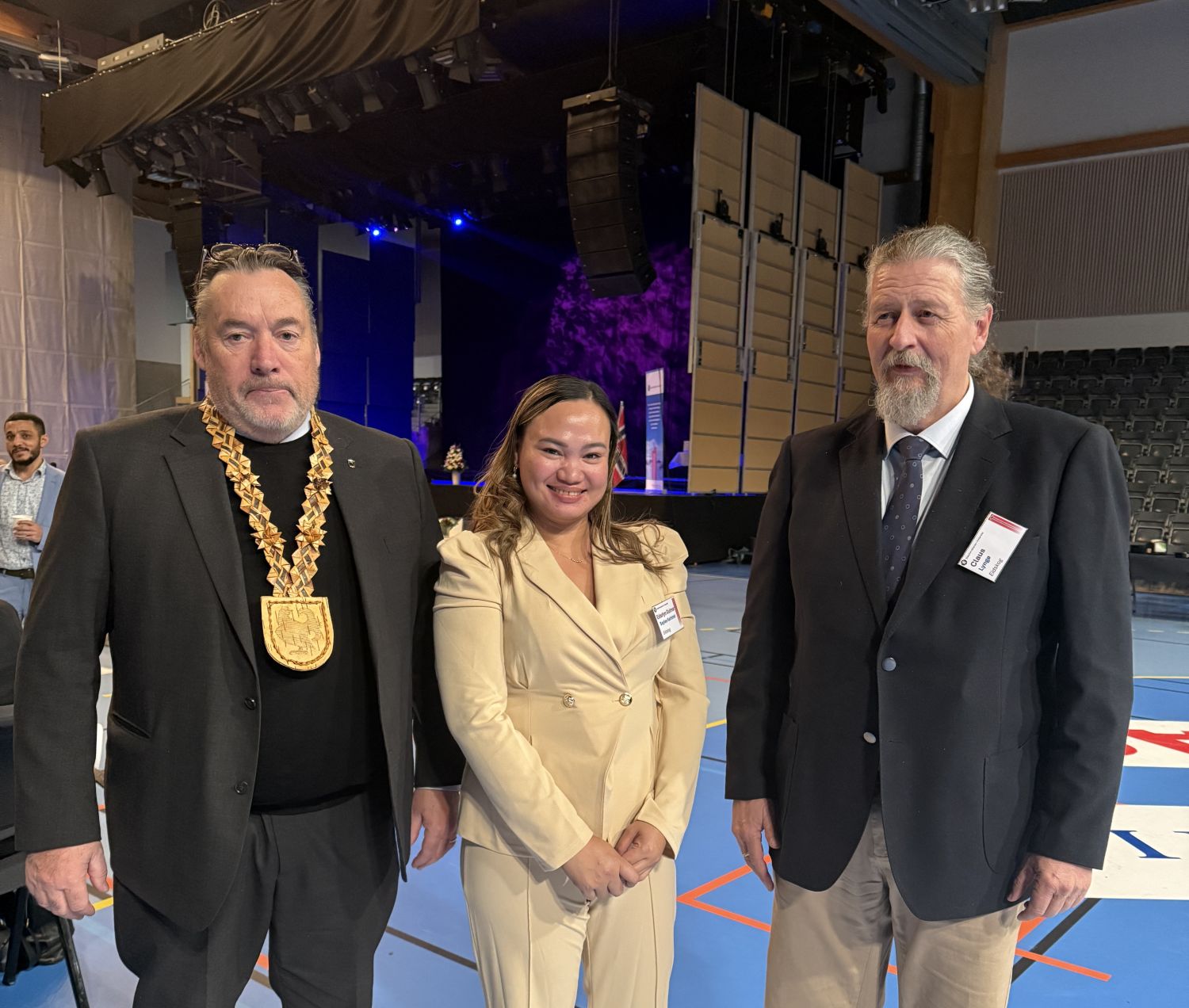 En kvinne og to menn fra eidskog står oppstilt i en festpyntet hall. 