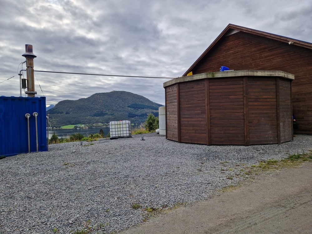 Biogasskum inntil hus, begge kledd i brunbeisa treverk. Utsikt mot fjell i bakgrunnen.