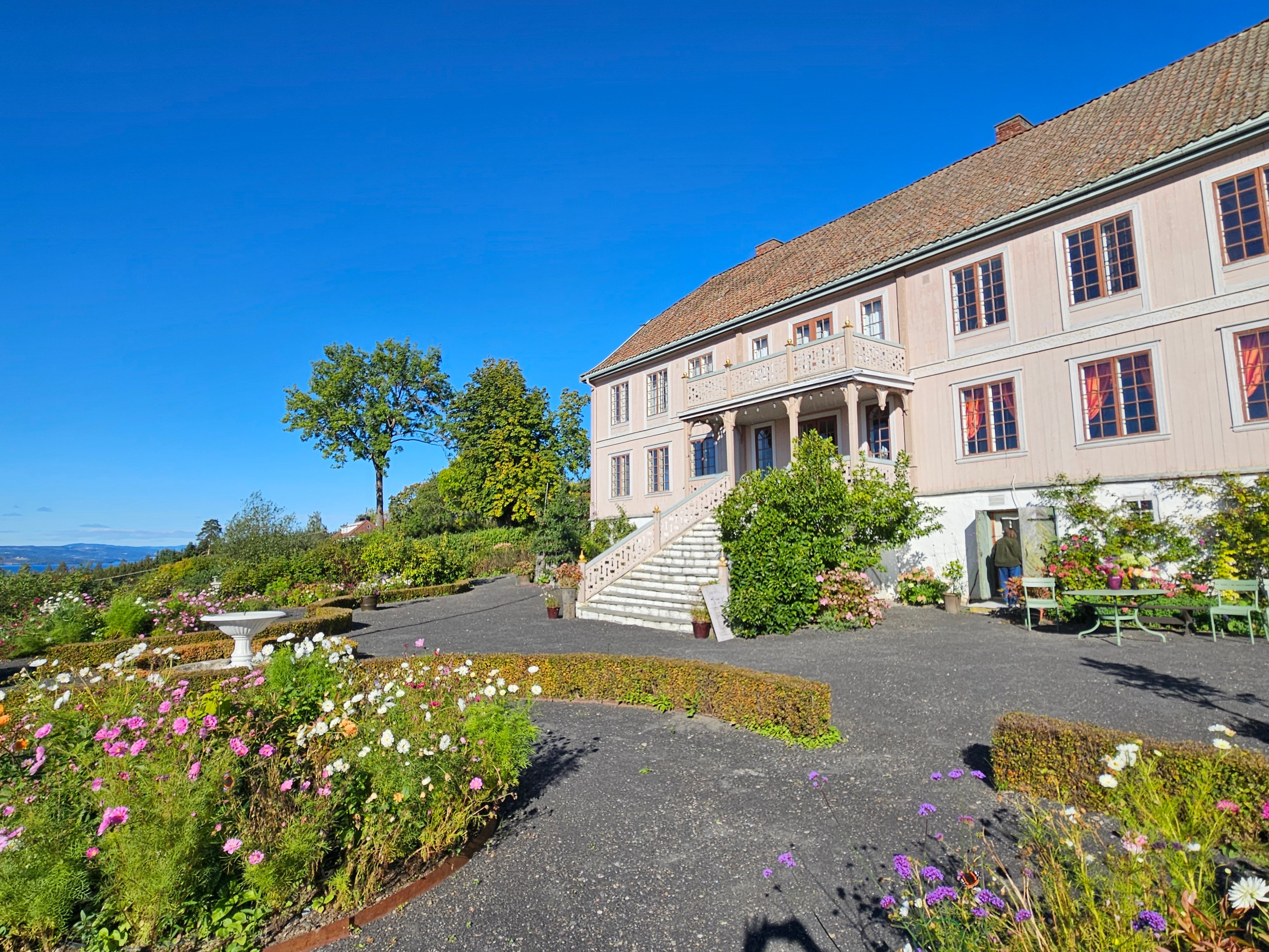 Hageanlegg med mørke grusganger og frodige blomsterbed foran en stor, blekrosa hovedbygning med brei trapp ned i hagen.