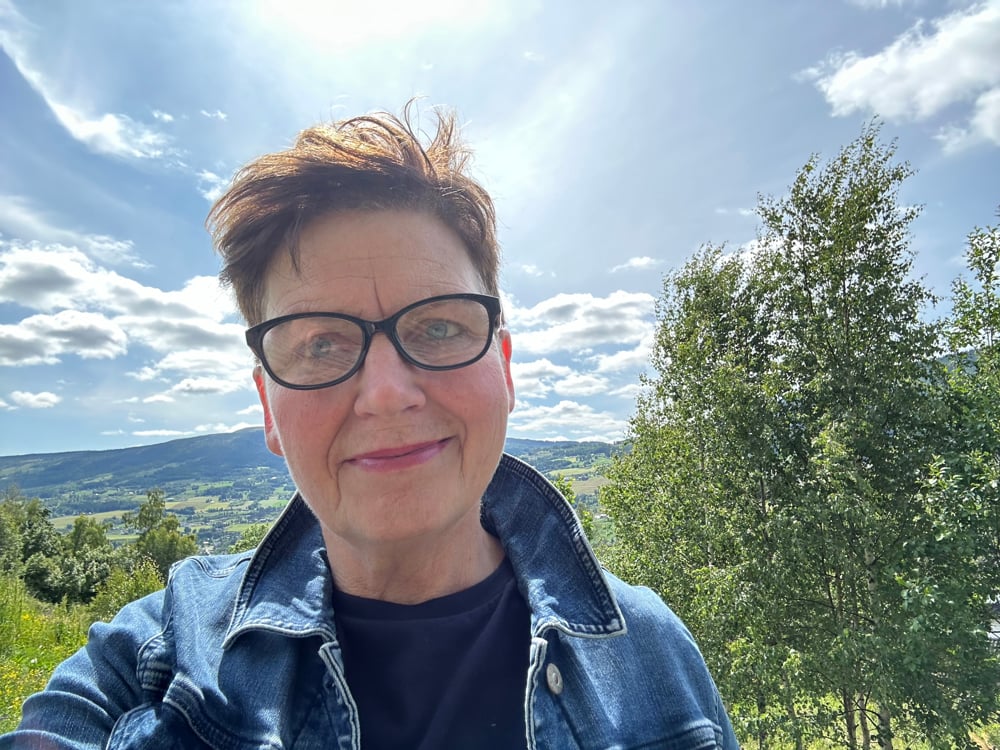 Kvinne kledd i dongeri jakke. Bjørketre og blå himmel bak.