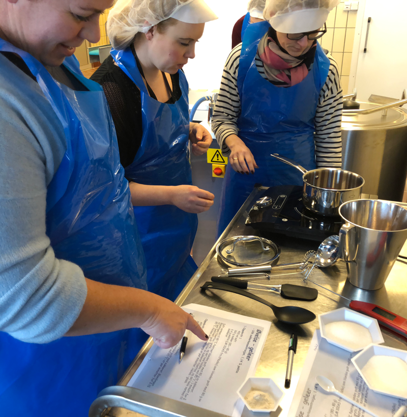Foto: Kompetansenettverket Lokalmat. Tre personar med fokle og hetter på hovudet, ved ein kjøkenbenk der ulike ingrediensar er målt opp og ulik reiskap ligg framme.