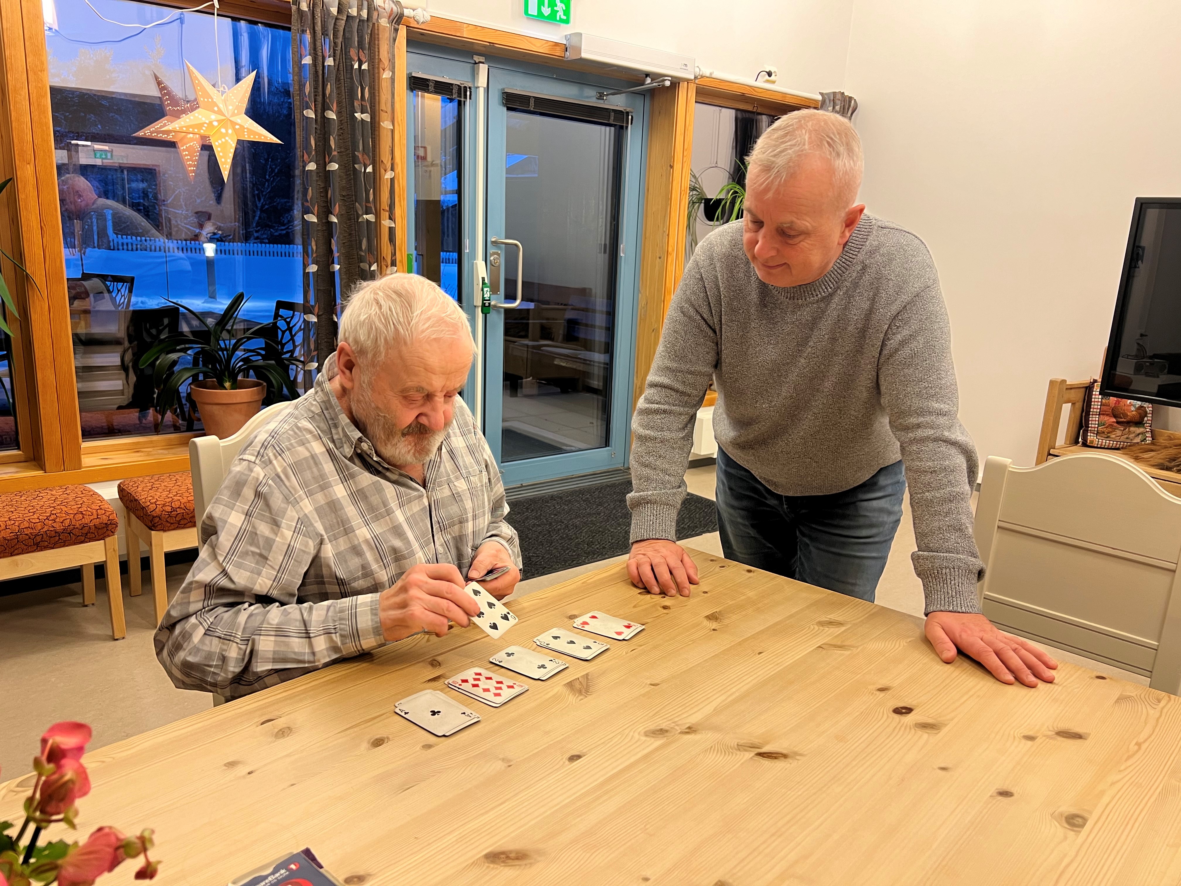 Per Bruimellom i hyggelig prat med Knut Storberget på Skjåkheimen. Foto: Edel Kveen. To eldre menn snakker sammen ved et bord. Den ene legger kabal.